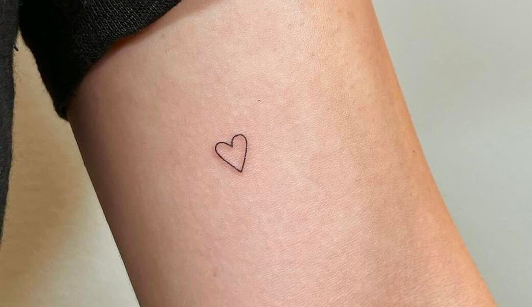 A very small outlined heart tattooed on an arm, drawn in one continuous hairline stroke.